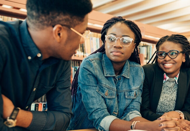 Three young Black students lean over a table in a library. Two women with glasses appear to be listening to their male colleague whose head is turned away from the camera and towards them.